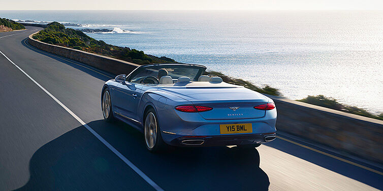 Bentley Edinburgh New Bentley Continental GTC Azure convertible rear view in Light Windsor blue paint with 22 inch ten spoke wheels driving dynamically on an ocean road in the sunshine