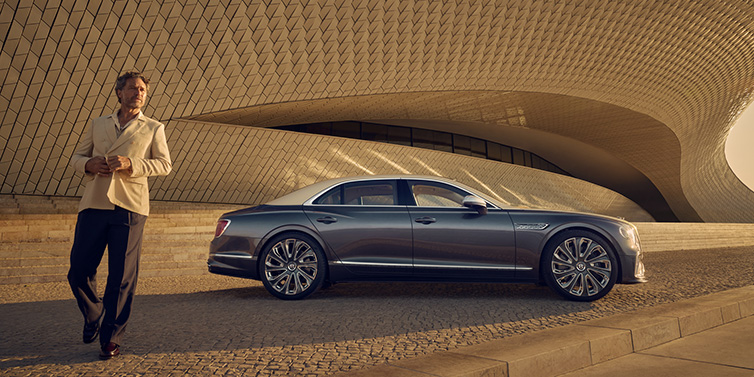 Bentley Edinburgh Bentley Flying Spur Mulliner sedan in Rose Gold over Magnetic duotone paint parked by a curved wall with person in suit jacket standing on the left